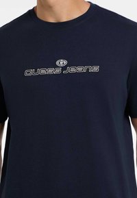 Tumedat sinist puuvillane T-särk, millel on ümar kaelus ja lühikesed varrukad. Rinnal on valge ja halli värvi tikitud "Guess Jeans" logo.