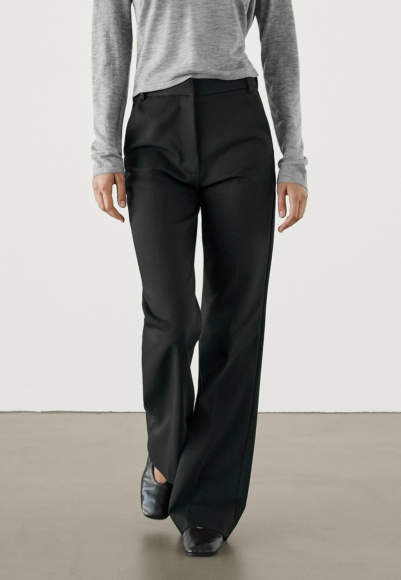 Pantalons larges noirs en tissu lisse, présentant une coupe ajustée et un devant plat. Portés avec un haut gris à manches longues et des chaussures noires.