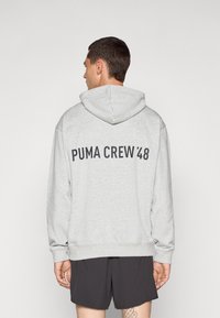 Siva majica s kapuljačom na vezanje, s natpisom "PUMA CREW '48" u crnoj boji na leđima. Napravljena od mekanog, teksturiranog materijala.