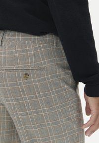 Spodnie w beżowo-szary wzór w kratę, z tylną kieszenią z zapięciem na guzik i dopasowanym krojem. Wykonane z teksturowanej tkaniny.