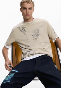 Beige shirt with bird and fish designs, short sleeves, layered over a white top; paired with dark denim jeans featuring a patch detail.