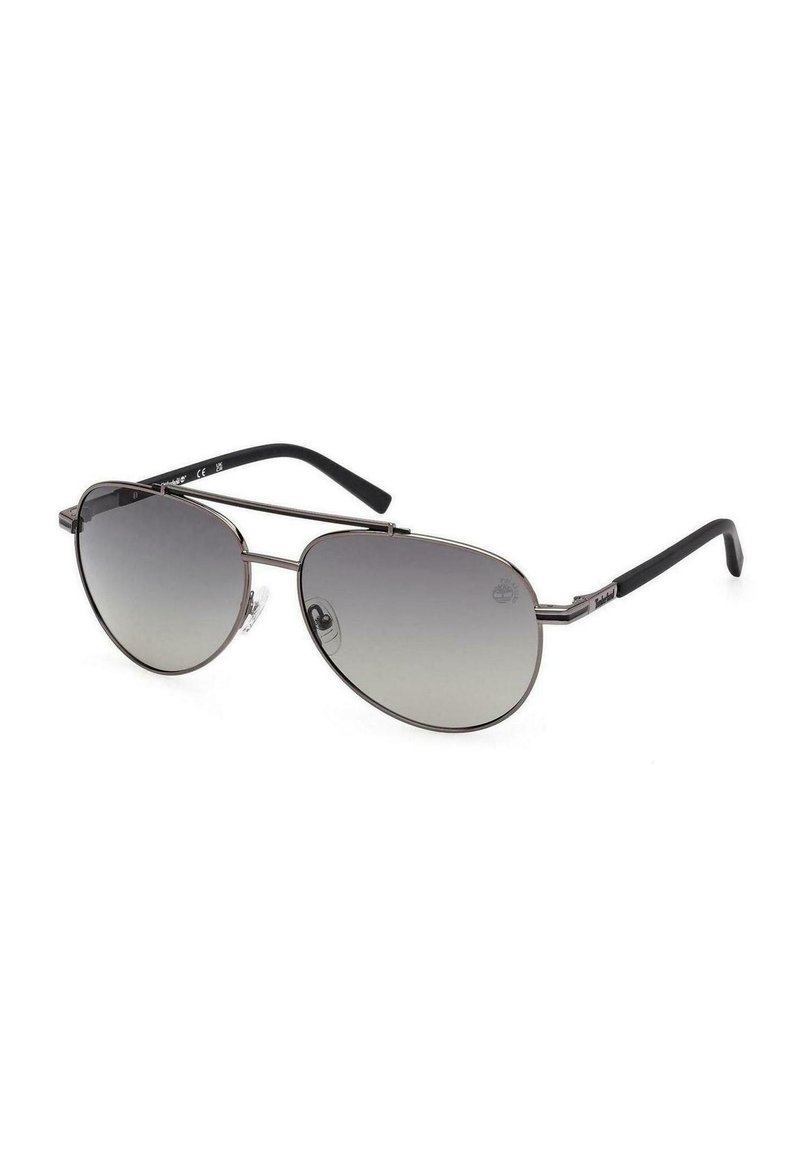 Gafas de sol tipo aviador con finos marcos de metal plateado, lentes grises degradados y puntas de las patillas negras, reposando sobre una superficie blanca.