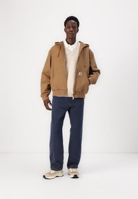 Veste à capuche brune avec poches avant, superposée sur un léger pull et une chemise blanche. Assortie avec un jean bleu et des baskets beiges.