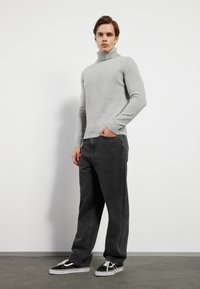 Grå turtleneck genser laget av mykt stoff, parret med mørkegrå jeans og svarte joggesko, stående pose med hendene i lommene.