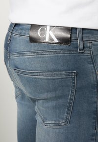 Bleka blå denimjeans med en synlig svart etikett som säger "Calvin Klein Jeans" på baksidan, med standardfickor och sömnadsdetaljer.