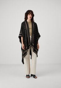 Zwarte poncho met een textuurpatroon, gouden accenten en franjes; gecombineerd met een beige top en lichtgekleurde wijde broek.