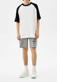 Henkilö, joka on pukeutunut valkoiseen ja mustaan lyhythihaiseen raglan-paitaan, revittyihin vaaleansinihin farkkushortseihin, valkoisiin sukkiin ja valkoisiin tennareihin, seisoo valkoista taustaa vasten.