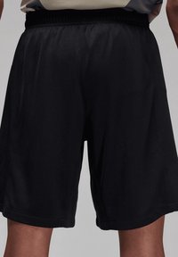 Shorts de sport noirs longueur genou portés par une personne, avec une taille élastique et une coupe ample sur un fond uni.