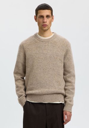 Homme portant un pull en laine beige et un pantalon marron foncé, debout avec une main dans la poche sur un fond uni.
