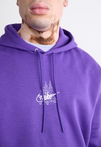 Lilla hettegenser laget av myk stoff, med hvit brodert grafikk og snorer. Designet inkluderer en naturscene og tekst.
