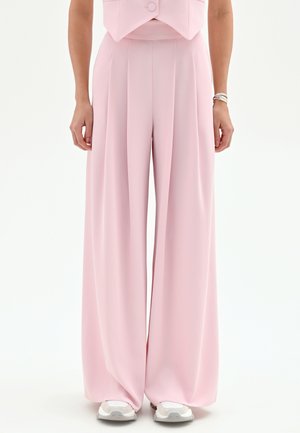 Donna che indossa pantaloni rosa pallido a vita alta e gamba larga con pieghe, abbinati a sneaker bianche e un top rosa corto.