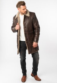 Abrigo de cuero marrón con cuello de borrego, cierre frontal de botones y bolsillos laterales. Lleva puesto sobre una camisa blanca y jeans oscuros, mostrando un estilo casual.