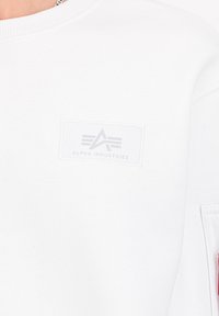 Bela sweatshirt iz mehkega blaga, z teksturirano logotipno nalepko na prsih, ki vsebuje besedilo "ALPHA INDUSTRIES" in dekorativno šivanje.
