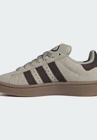 Sneaker en daim gris avec triples bandes marron foncé, lacets assortis et semelle marron, vu de profil sur un fond clair.