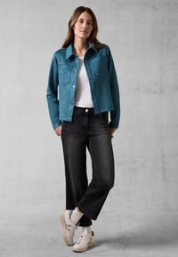 Tealfärgad mockajacka med framfickor, bärs över en vit T-shirt och svarta vida byxor, i kombination med vita sneakers och synliga strumpor.