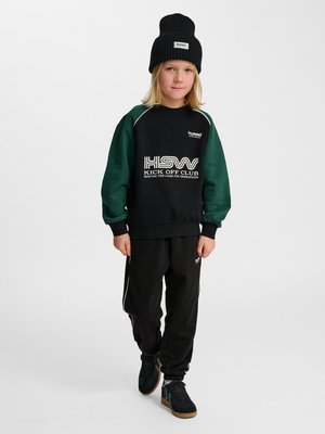 Kind trägt schwarze Mütze, schwarzen und grünen Sweatshirt mit dem Schriftzug „KICK OFF CLUB“, schwarze Hose und schwarze Sneakers, steht vor weißem Hintergrund.