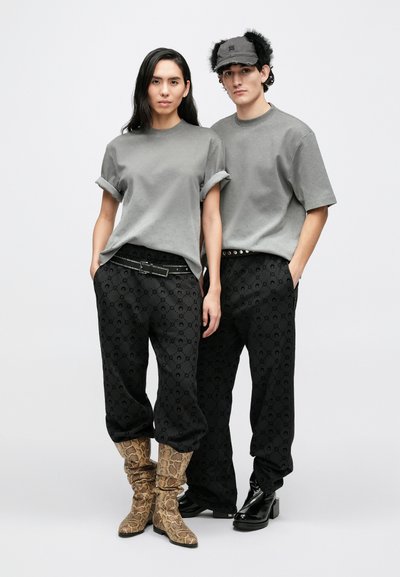 Dwoje modeli nosi dopasowane, szare, oversize'owe t-shirty z podwiniętymi rękawami, zestawione z czarnymi, wzorzystymi spodniami. Kobieta ma na sobie botki w wężowy wzór, a mężczyzna błyszczące, czarne buty.