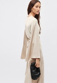 Smėlio spalvos satino sijonas su plačiomis rankovėmis ir rišamu juosmeniu, laikomas juodos odos delninuke. Lygus tekstūra, minimalistinis dizainas.