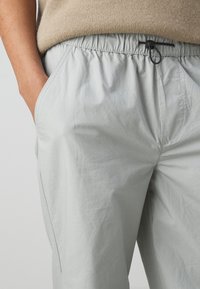 Personne portant un pantalon gris clair avec une taille élastique et un cordon noir, main placée dans la poche gauche, haut beige visible au-dessus de la taille.