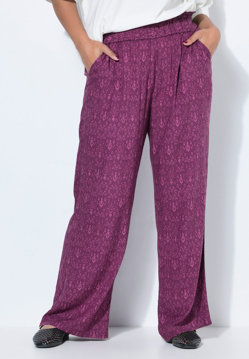 Volné fialové kalhoty s vzorem a elastickým pasem, nošené s černými slip-on botami a rukama v kapsách.