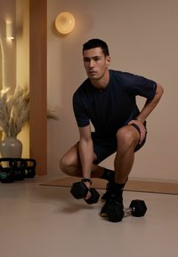 Homme en tenue de sport noire accroupi en train de soulever un haltère dans une salle d'entraînement minimaliste avec des kettlebells et des plantes séchées à proximité.