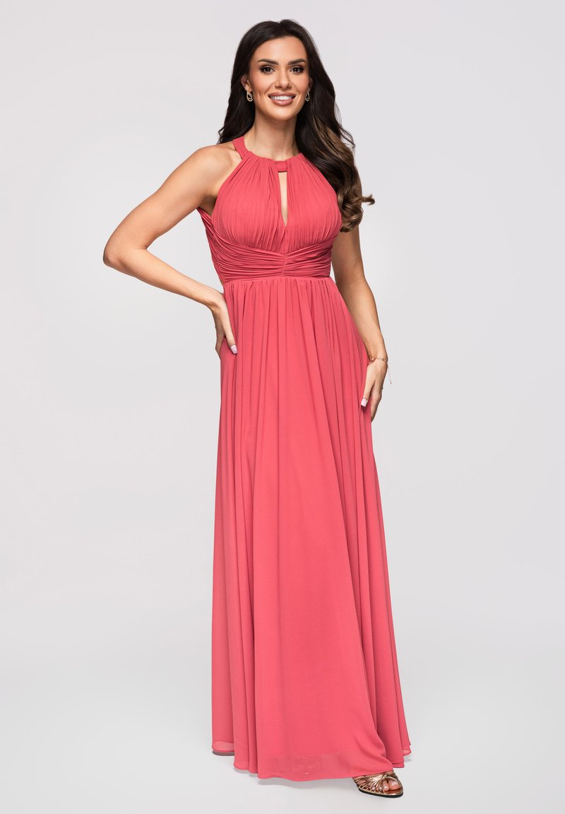 Femme aux longs cheveux foncés portant une robe corail plissée sans manches à col halter, debout avec une main sur la taille et souriante.
