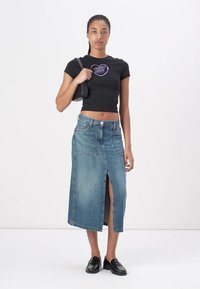 Svart cropped t-skjorte med et lilla grafisk design, kombinert med et blått denimskjørt med splitt foran, og svarte sko. En enkel, uformell design.