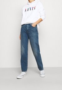 Persona vistiendo una sudadera blanca de Levi's con un logo colorido, jeans rectos azules y zapatillas de plataforma blancas, de pie contra una pared sencilla.