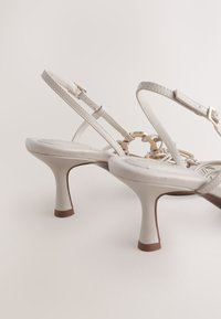 Sandales à talons en cuir blanc avec une fine bride à la cheville, un accent de chaîne dorée et un talon stiletto de hauteur moyenne ; texture lisse et design minimaliste.