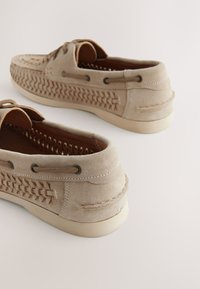 Chaussures bateau en daim beige clair avec un motif tissé sur le côté, des lacets marron, des accents texturés et une semelle en caoutchouc de couleur crème.