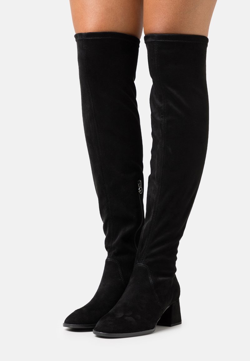 Bottes noires en daim au-dessus du genou avec talons carrés et fermetures éclair latérales portées par une personne aux jambes nues sur un fond uni.