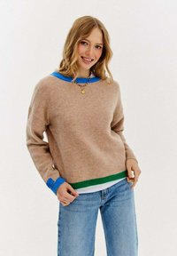 Beżowy sweter dzianinowy z niebieskimi i zielonymi ściągaczami przy mankietach i u dołu. Posiada luźny krój oraz okrągły dekolt, noszony na jasnoniebieskich dżinsach.