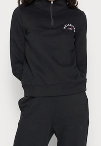 Svart zip-up sweatshirt med högt krage, med en liten rosa "Hollister"-logotyp. Mjuk tyg med ribbad mudd och nederkant.