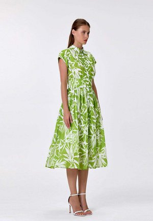 Femme portant une robe midi verte sans manches avec des imprimés botaniques blancs et des sandales blanches à talons hauts, debout devant un fond uni.