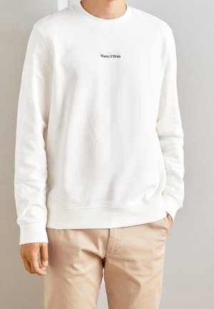 Person iført en hvid langærmet sweatshirt med teksten "Marc O'Polo" og beige bukser, stående mod en ensfarvet lysefarvet væg.