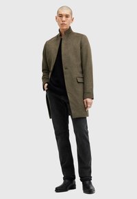 Cappotto in misto lana verde oliva con colletto alto, chiusura a un bottone, tasche laterali, abbinato a jeans neri e stivali alla caviglia.