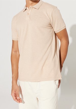 Kort beige poloshirt med tekstureret stof, tre-knaps stolpelukning og et lille emblem på brystet. Kombineret med lyse bukser.