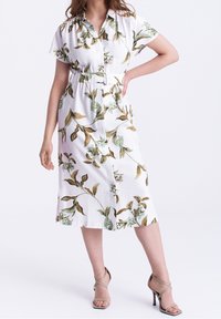 Donna che indossa un abito midi bianco con stampa floreale verde e marrone, vita cintrata, maniche corte e sandali con tacco alto beige a strappi.