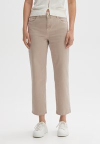 Mujer vistiendo pantalones rectos beige, zapatillas blancas y un suéter corto blanco texturizado contra un fondo blanco liso.