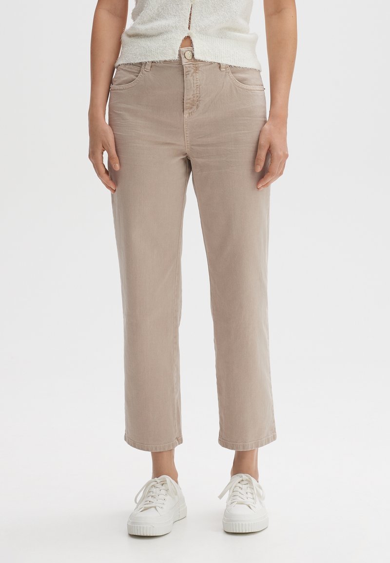 Mujer vistiendo pantalones rectos beige, zapatillas blancas y un suéter corto blanco texturizado contra un fondo blanco liso.