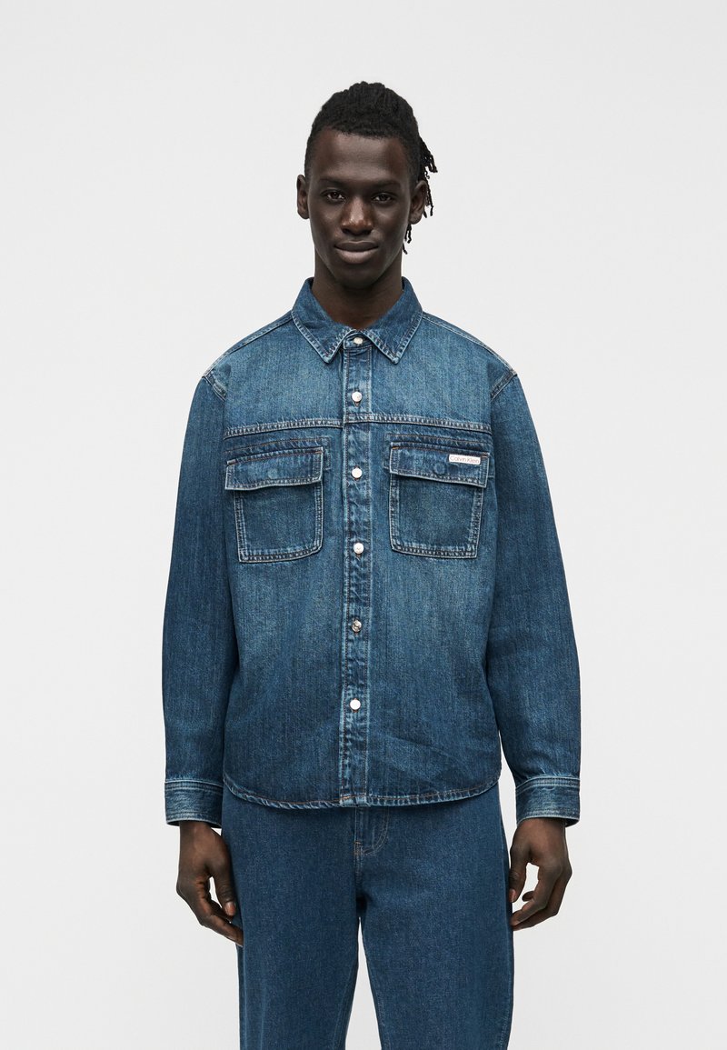 Άνδρας με σκούρο δέρμα που φορά ένα κουμπωμένο τζιν μπουφάν με δύο τσέπες στο στήθος και ταιριαστό τζιν παντελόνι, στέκεται μπροστά σε απλό φόντο.