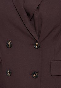 Blazer marron foncé croisé avec quatre boutons noirs et beige marbrés et un col à revers cranté.