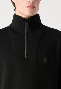 Juodas zip-up megztinis su aukštu apykakle, tekstūruotas mezginys, vertikali auksinė užtrauktuko sistemos dalis ir apvalus logotipo pleistras ant krūtinės.