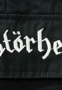Svart stoff med hvit brodert tekst, med stilisert skrifttype. Teksten viser lett slitasje. Bakgrunnen er teksturert med synlig søm.