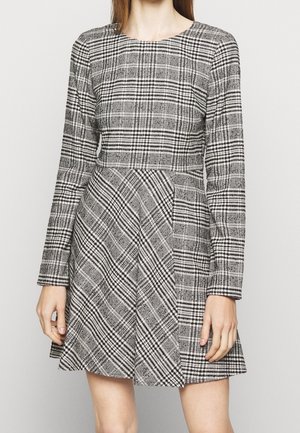 Mujer con un vestido de mangas largas de cuadros en blanco y negro, con cintura ajustada y falda acampanada, de pie frente a un fondo liso.