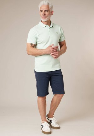 Homme âgé aux cheveux et barbe blancs, portant un polo vert clair, un short bleu marine, des baskets blanches, debout les mains croisées, regardant sur le côté.