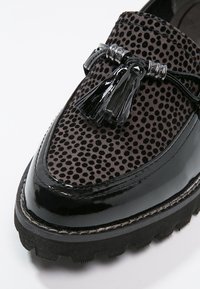 Mocassin noir avec garniture en cuir verni brillant, dessus en tissu texturé à pois, pompons décoratifs et semelle épaisse à crampons.