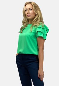 Zielona bluzka z krótkim rękawem, z warstwowymi falbanami i gładką fakturą. Połączona z ciemnoniebieskimi dopasowanymi spodniami z bocznymi kieszeniami.