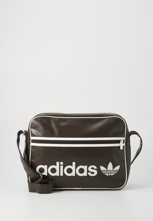 Mørkebrunt rektangulært Adidas skuldertaske med hvidt logo, hvid lynlås på den forreste lomme og justerbar rem mod hvid baggrund.