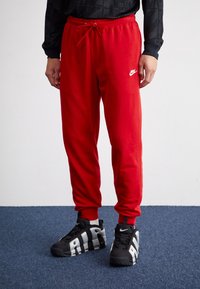 Pantalons de survêtement rouges avec taille élastique et cordon, arborant un logo Nike blanc. Associés à des baskets noires avec des accents gris audacieux.